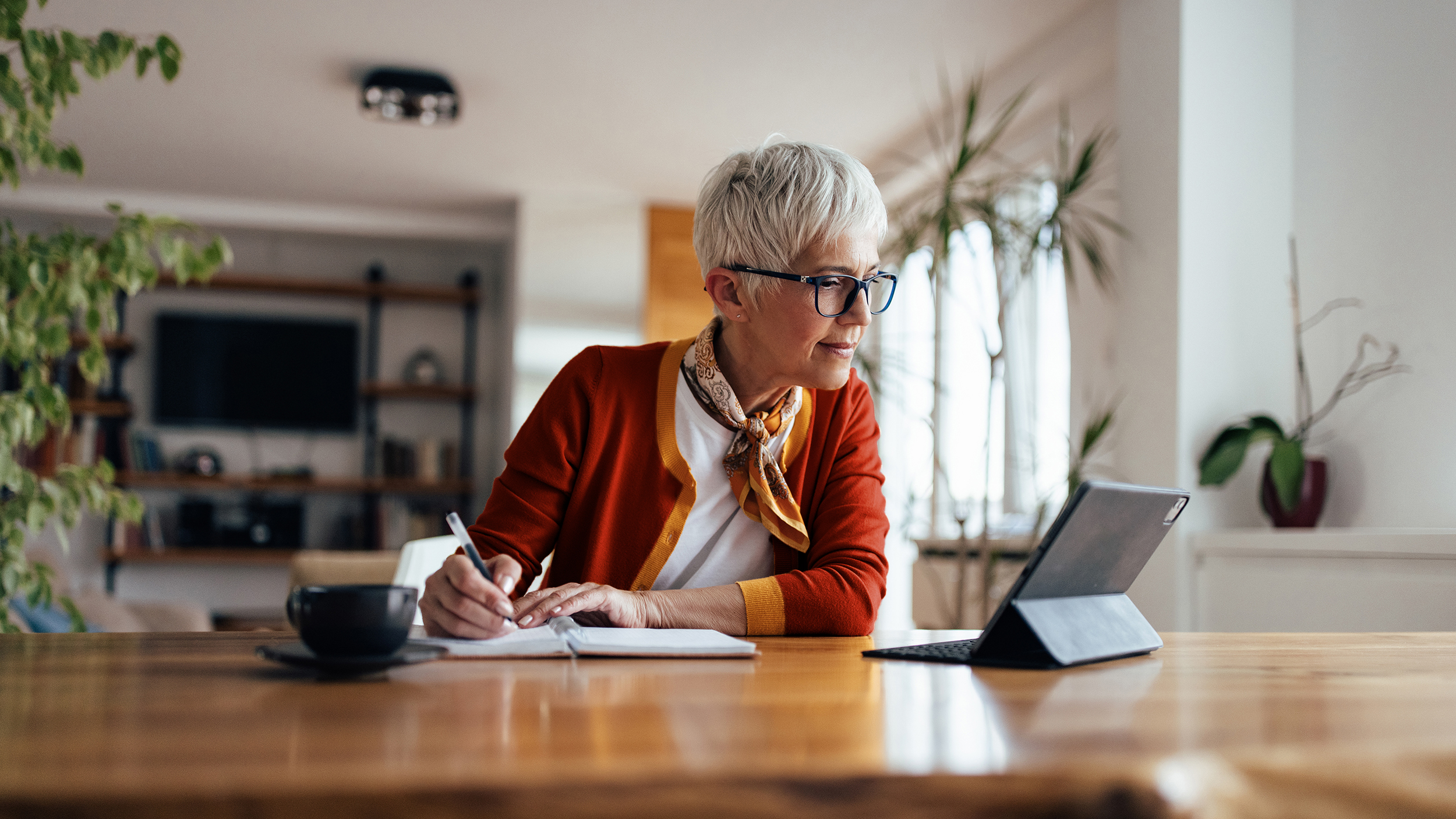 A stylish older woman looking at a computer screen and writing something down. 